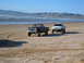 1985 Ford Ranger - Two pickup trucks drive on a sandy beach near the ocean with mountains in the distance.