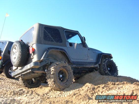 Gray Jeep Wrangler with large tires climbing a sand dune, another vehicle partially visible to the left