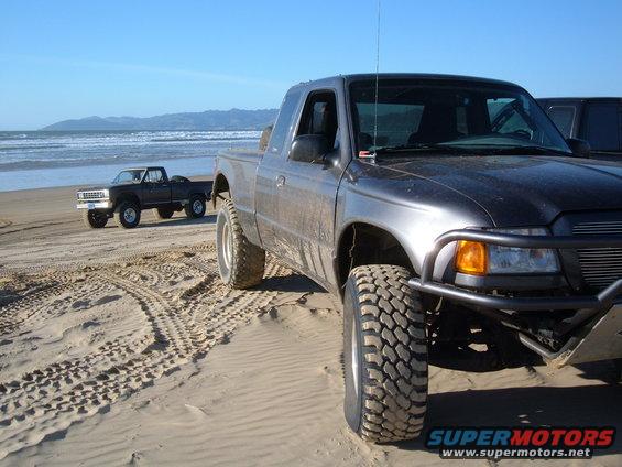 Gray pickup truck with off-road tires on sand, front bumper visible. Another dark pickup truck in the background on the beach