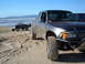 1985 Ford Ranger - Two pickup trucks are parked on a sandy beach with the ocean and hills in the background.