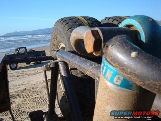 Close-up of off-road suspension with a spare tire. A black pickup truck is visible on the beach in the background
