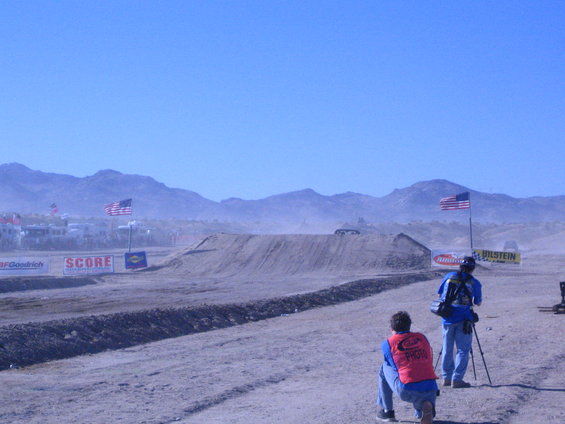 Dirt race track with two photographers in foreground, American flags, and mountains in background