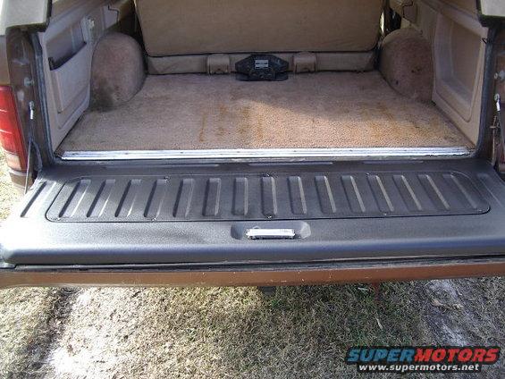 Interior cargo space of a brown SUV, showing carpeted floor and seats folded up. A black electronic device sits on the floor