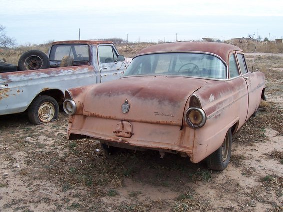 1955 Ford Customline sedan parked next to a pickup truck, both showing rust