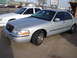 2000 Ford Crown Victoria - Two sedans, a silver and a white one, parked on a paved lot.