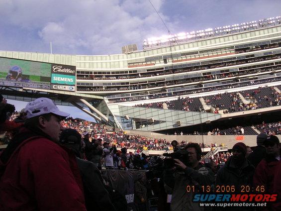 People in a stadium watching a game, with a large screen showing ads and a football player