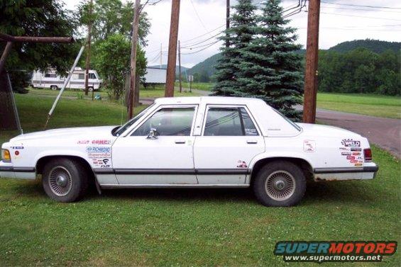 1988 Mercury Grand Marquis sedan with numerous stickers on the doors and rear quarter panel, parked on grass