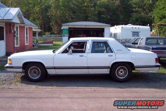 White sedan parked outside a garage with a trailer and another car behind it