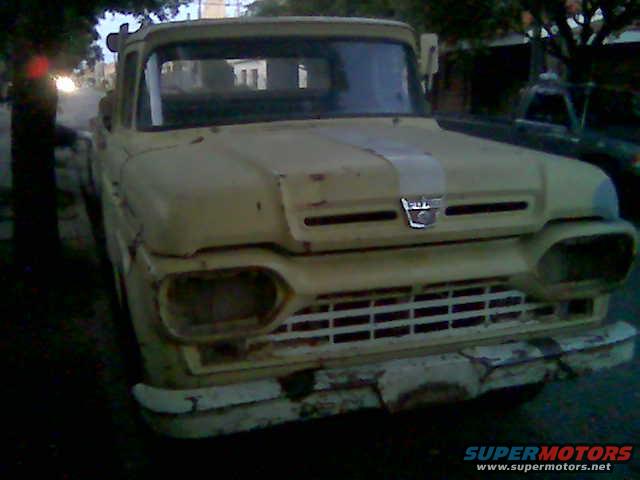 Front view of a yellow 1957-1959 Ford F-100 truck with missing headlights and a damaged bumper