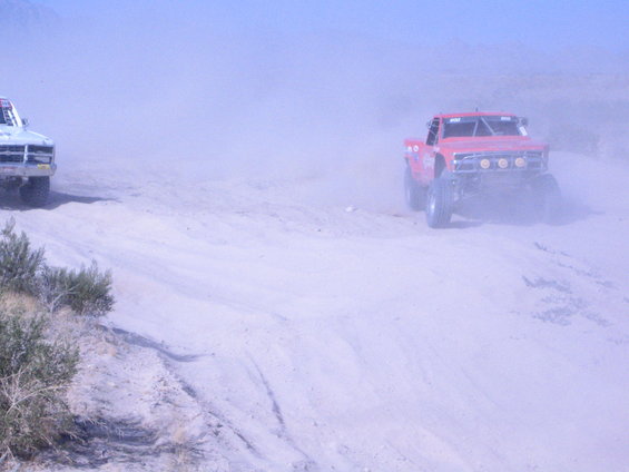 Two off-road trucks race through dust on a dirt track. One is red, the other is white