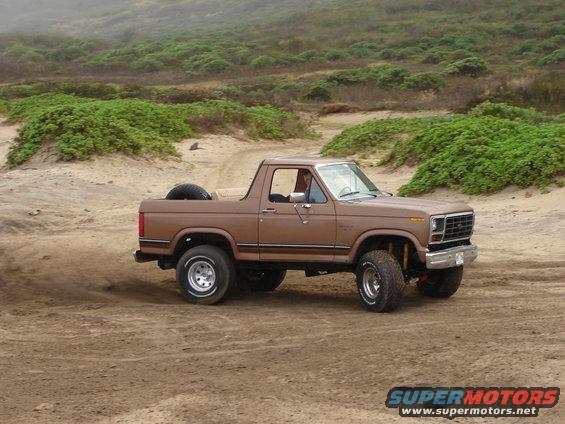 Brown 1985 Ford Bronco driving on sand, spare tire in the bed. Coastal hills in background