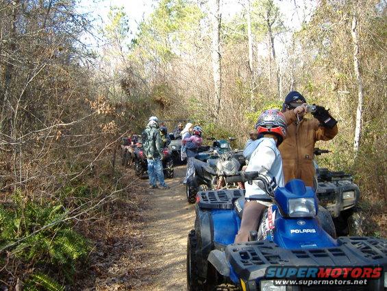 Group of people on ATVs lined up on a dirt path in a wooded area. One person is taking a photo