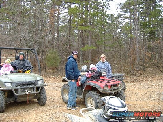 Red ATV with a person standing next to it, and a green utility vehicle with people inside, in a wooded area