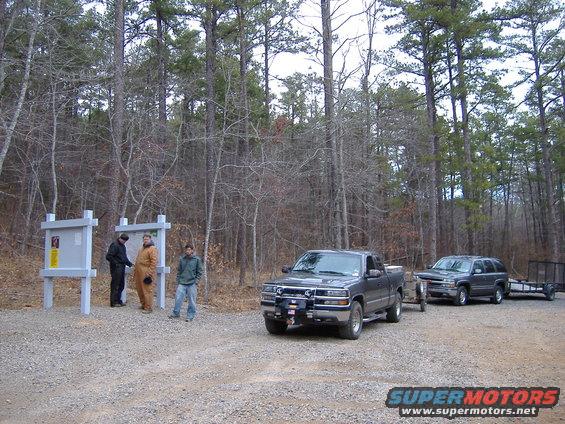 Two trucks and a trailer parked near a bulletin board in a wooded area