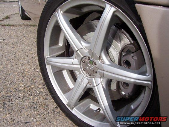 Close-up of a silver five-spoke aftermarket wheel with a drilled brake rotor and caliper. The vehicle body is beige