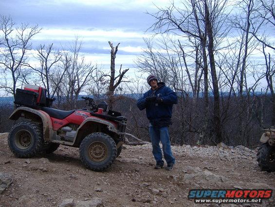 Red ATV with black storage box on rear, parked on dirt trail. A person stands beside it