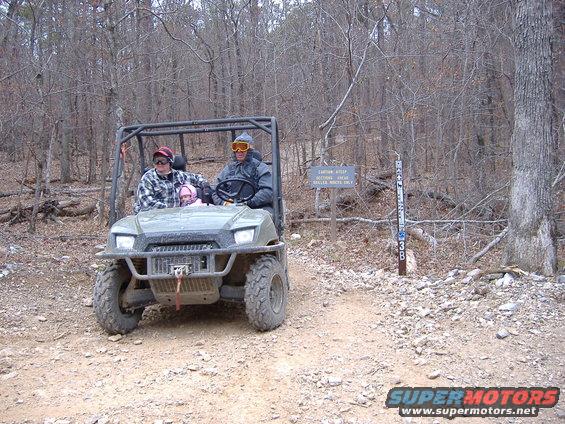 People riding in a utility vehicle on a dirt path in the woods. There's a sign that says 'CAUTION: STAY ON MARKED TRAILS. TRAILER HITCHES ONLY.'