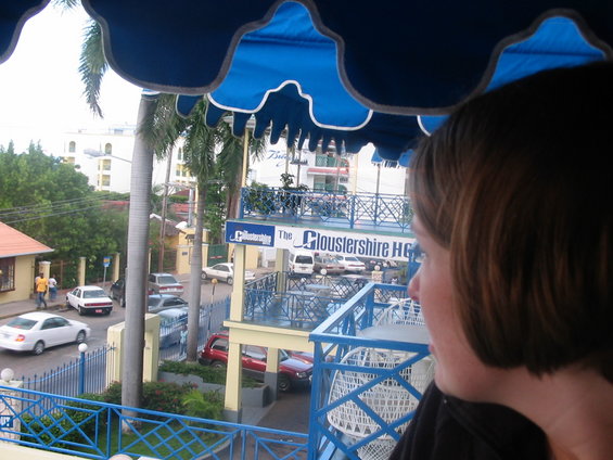 Street view from a balcony with several cars parked and driving, palm trees, and buildings. A woman's head is in the foreground
