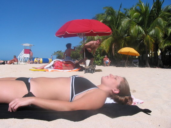 People sunbathing on a sandy beach with palm trees and umbrellas under a clear blue sky