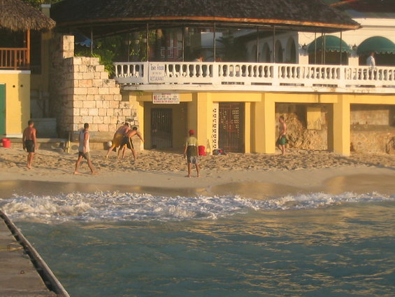 People playing volleyball on a sandy beach in front of a yellow building with a thatched roof structure
