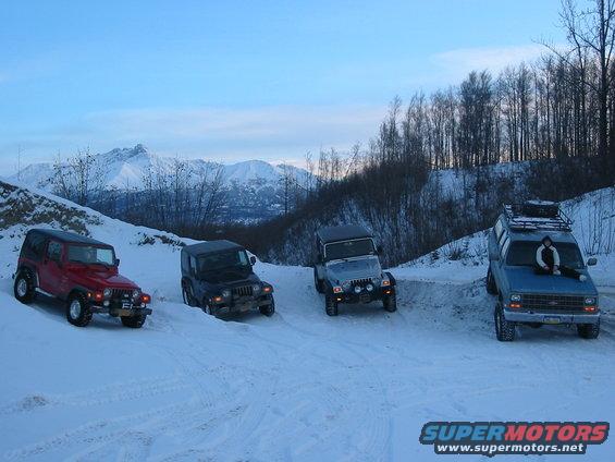 Four vehicles, three Jeeps and a van, parked on a snowy road with mountains in the background