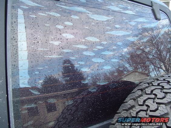 Rear window of a vehicle covered in water droplets and some light streaks, with a spare tire visible on the right