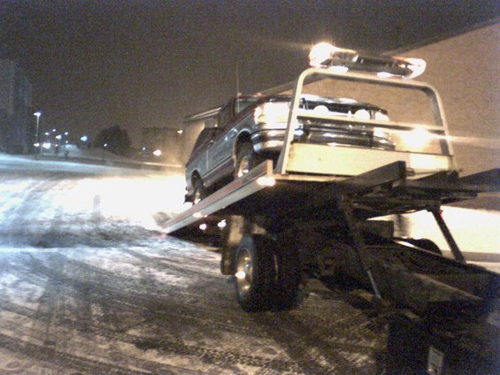 Tow truck carrying a dark-colored SUV at night in the snow
