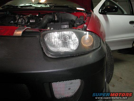 Front end of a red car with hood open, showing engine and headlight. White car parked next to it