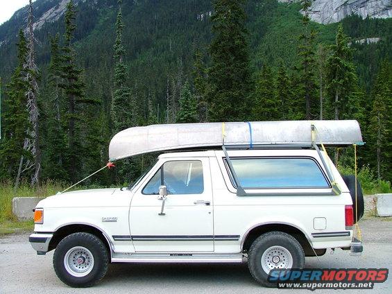 White Ford Bronco parked on a dirt road with a canoe on its roof rack, mountains and trees in the background
