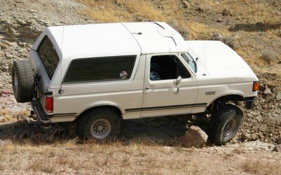 White Ford Bronco with camper shell and rear-mounted spare tire, angled right, driving on rocky terrain