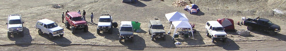 Several off-road SUVs and a pickup truck parked in a dusty, open area with tents and canopies