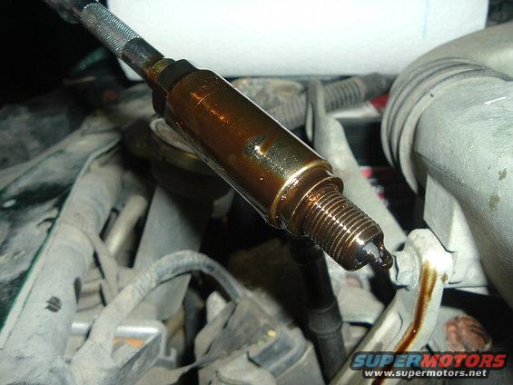 Spark plug covered in dark oil, held by a tool, with engine components visible