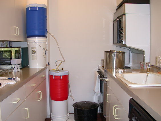 Kitchen counter with stacked buckets and brewing equipment, including a blue cooler on top of white buckets, connected by tubing to a red cooler