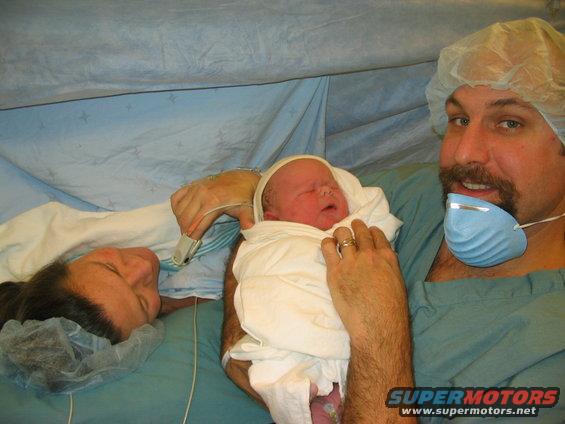 Newborn baby wrapped in white blanket held by father, mother looks on. Hospital setting