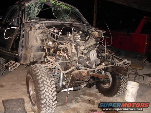 Wrecked SUV, front end removed showing engine and suspension. Cracked windshield has green text. Tow truck visible in background
