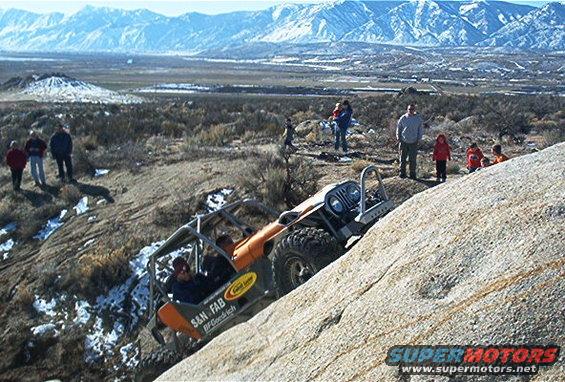 Off-road buggy with roll cage climbing a steep rock face, people watching on the hill
