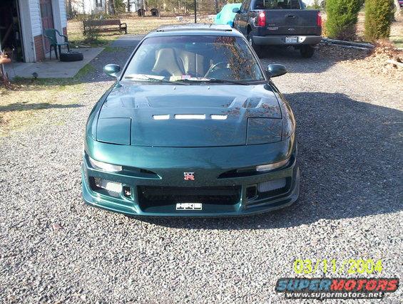 Green coupe with aftermarket body kit and a Nissan GT-R badge on the grille. A black pickup truck is parked behind it