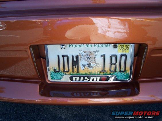 Rear of an orange car showing a license plate with a panther graphic and 'JDM 180' text. Also has a 'nismo' badge
