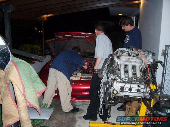 Three guys working on a red car with the hood up, and a V6 engine on a stand nearby