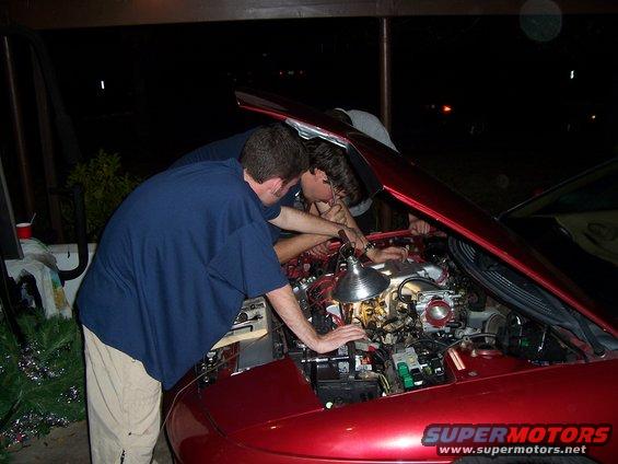 Red car engine bay with three people working on the engine under a lamp