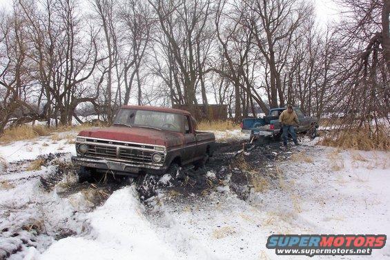 Red pickup truck stuck in mud and snow, another truck behind it with a person standing nearby
