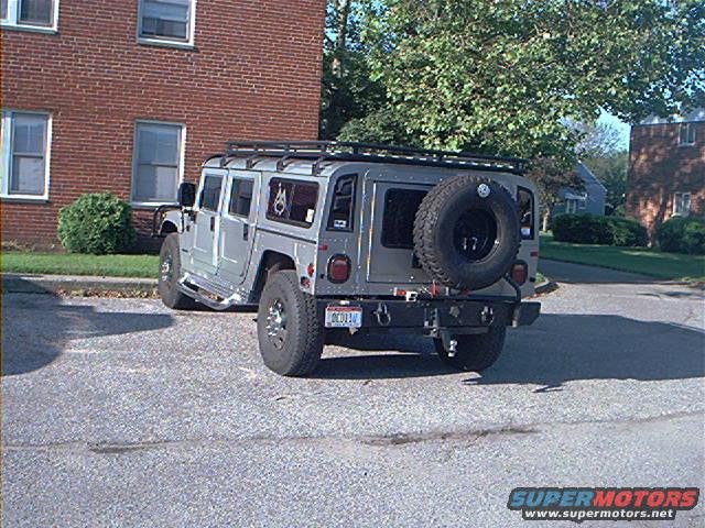 Silver Hummer H1 with roof rack and spare tire on rear, parked on asphalt