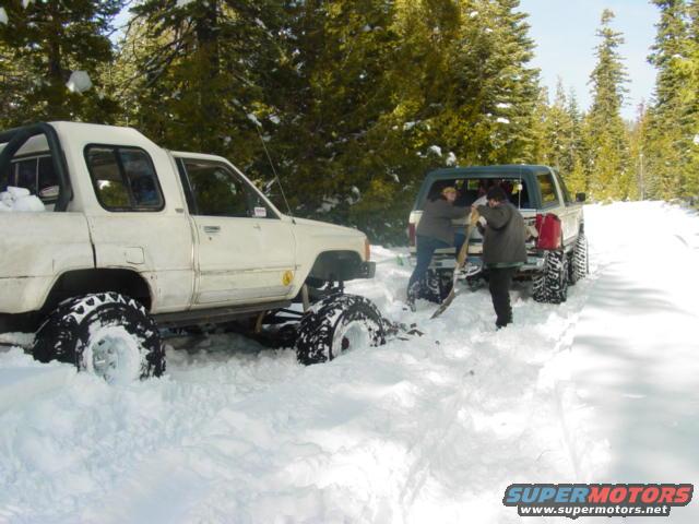 White pickup truck with big tires stuck in snow next to another truck where people are digging it out