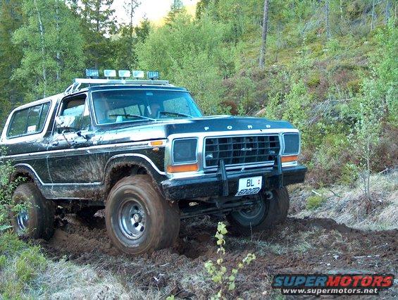 Black Ford Bronco driving through mud on a wooded trail. It has a roof rack with lights