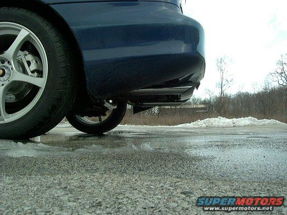 Rear bumper and wheel of a dark blue car, parked on a wet, icy road with snow piles in the background