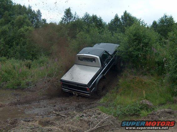 Black pickup truck with diamond plate bed cover stuck in mud off-road. Trees and bushes surround the muddy track