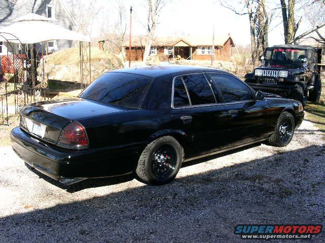 Black 2001 Ford Crown Victoria sedan parked next to a black Jeep, viewed from the rear three-quarter angle