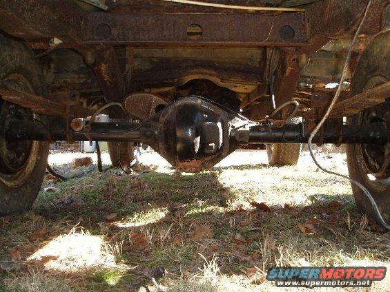 Underside view of a vehicle's rear axle, differential, leaf springs, and tires. The frame and undercarriage show significant rust