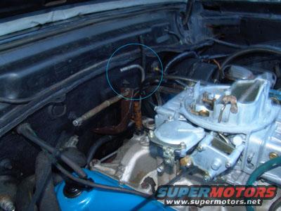 Engine bay showing a carburetor, brake booster, and various hoses. A blue circle highlights a metal line