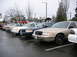 1999 Ford Crown Victoria - Multiple police sedans, including a tan Ford Crown Victoria, are parked in a lot on a cloudy day.